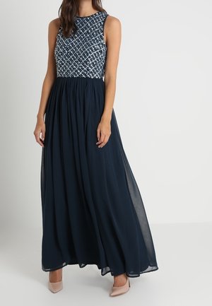 Woman wearing sleeveless navy blue gown with embellished patterned bodice and flowing skirt, paired with nude pointed heels.