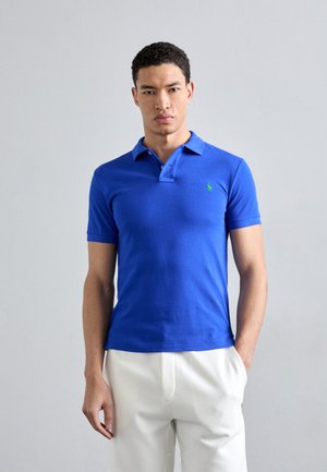 Polo shirt blu con un logo verde sul petto, caratterizzato da un colletto classico e maniche corte, abbinato a pantaloni bianchi.