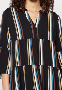 Černé šaty s vertikálními pruhy v modré, hnědé a bílé barvě. Mají výstřih do V, rozšířené rukávy a vícestupňový design. Měkká textura látky.