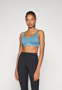 Sujetador deportivo azul claro con tirantes anchos y diseño sin costuras. El material es suave y elástico, con un logo minimalista en la parte delantera.