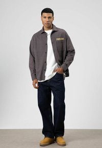 Geruite grijze flanellen blouse met de tekst "FREEDOM", witte T-shirt eronder, blauwe loszittende jeans en beige laarzen op een model.