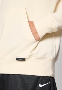 Světlá krémová mikina s kapucí, přední kapsou a žebrovaným lemem. Má štítek "CITY EDITION" a je doplněna texturovanými černými šortkami.