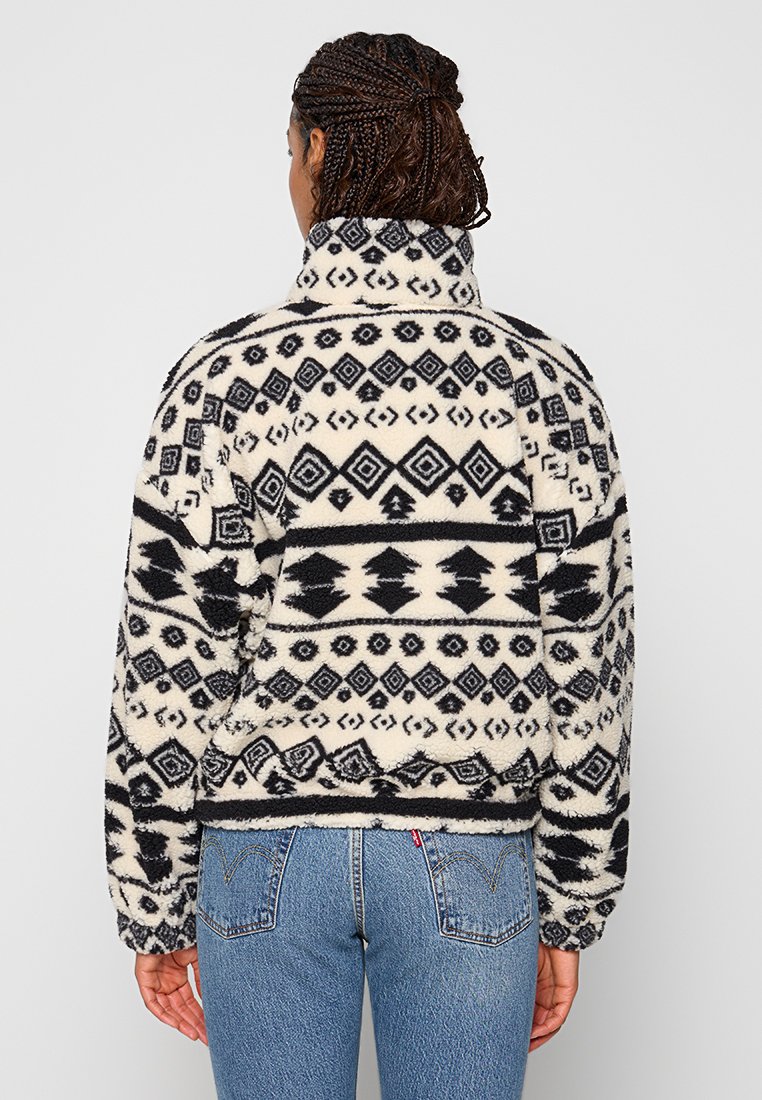 Fleece-Jacke mit hohem Kragen, geometrischen Mustern in Schwarz und Creme sowie einem entspannten Schnitt, kombiniert mit hochgeschnittenen Jeans.