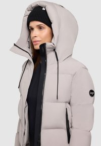 Pufferjakke i lys grå med en stor hætte, sorte detaljer og lynlås foran. Har et logo-patch på venstre arm.