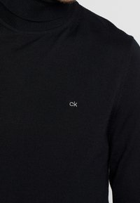 Maglione a collo alto nero a costine, realizzato in un tessuto lavorato a maglia, con un piccolo logo ricamato "ck" bianco sul petto.