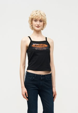 Młoda kobieta o krótkich, kręconych, blond włosach, ubrana w czarny krótki top z pomarańczowym napisem oraz ciemnoniebieskie jeansy, stojąca na jednolitym tle.