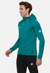 Mężczyzna w tealowej kurtce z kapturem na zamek i czarnych spodniach z logo, stojący bokiem na jednolitym tle.