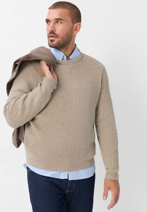 Homme barbu portant un pull côtelé beige sur une chemise bleue, tenant une veste marron sur l'épaule, regardant vers la gauche sur fond blanc.