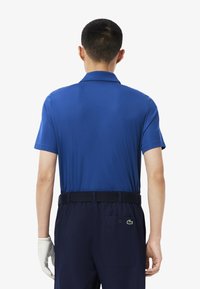 Korte mouwen polo shirt in felle blauwe stof, met een klassieke kraag, gecombineerd met marineblauwe broek en een witte handschoen zichtbaar op de linkerhand.