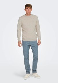 Suéter de punto beige claro con motas, cuello redondo, puños acanalados, combinado con jeans azul claro y zapatillas blancas. Diseño simple, corte casual.