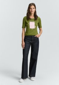 Groene T-shirt met korte mouwen, versierd met een roze grafiek en tekst, gecombineerd met donkerblauwe flared jeans en witte sneakers, staand op een neutrale achtergrond.