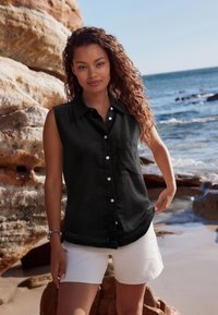 Schwarzes ärmelloses Hemd mit Knopfleiste, Brusttasche und seitlichen Spitzendetails, kombiniert mit hellen Shorts, an einem Strand.
