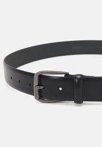 Ceinture en cuir noir avec une texture lisse, dotée d'une boucle en métal de couleur argentée. Ajustable avec plusieurs trous le long de la sangle.