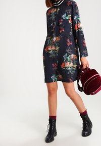 Sukienka w kwiatowy wzór w ciemnym granacie, z kołnierzykiem, długimi rękawami i przednią kieszenią. W zestawie z burgundowym plecakiem i czarnymi butami.