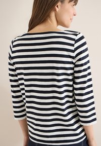 Femme de profil portant un t-shirt à rayures horizontales noires et blanches avec des manches trois-quarts, sur un fond uni.