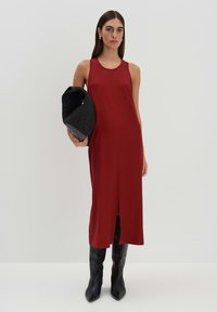 Rotes, ärmelloses Midikleid mit einem vorderen Schlitz, glatte Textur. Das Model hält eine schwarze, strukturierte Handtasche und trägt kniehohe schwarze Stiefel.