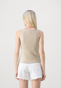 Kvinna med långt blont hår som bär en beige ärmlös topp och vita fransade denimshorts, sedd bakifrån mot en neutral bakgrund.