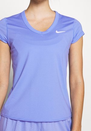Femme portant un haut de sport Nike à manches courtes en V de couleur violet clair et un short texturé assorti, sur un fond uni.