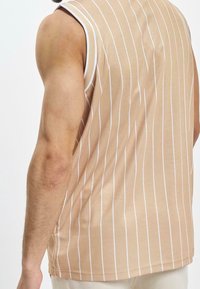 Beżowy, pionowo prążkowany bezrękawnik z teksturowanej tkaniny, z białymi paskami i okrągłym dekoltem, widziany z tyłu.