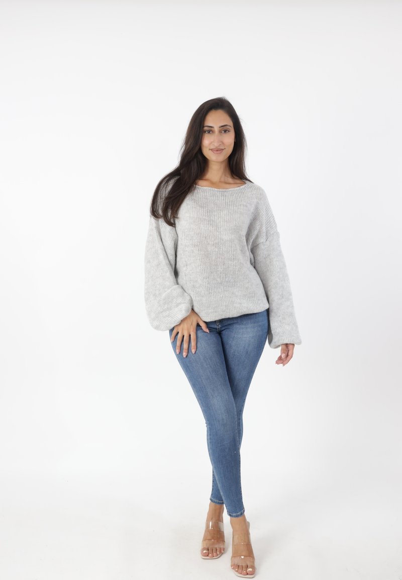 Lichtgrijze gebreide trui met brede mouwen, gecombineerd met aansluitende blauwe jeans en doorzichtige hakken. De trui heeft een brede halslijn en een gestructureerde afwerking.