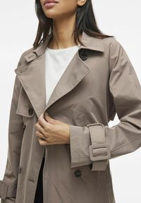 Trench coat di un leggero color taupe con design doppiopetto, ampio colletto, dettagli con bottoni e maniche con cintura. Tessuto liscio con vestibilità su misura.