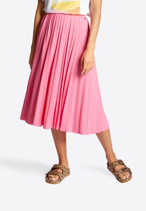 Femme portant une jupe plissée rose mi-longue, un t-shirt blanc avec un motif graphique, et des sandales marron à bride avec boucle et motif.