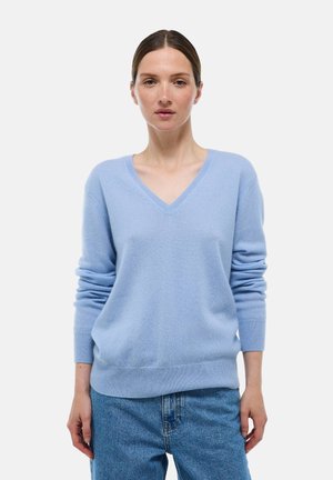 Femme aux cheveux bruns attachés, portant un pull léger bleu ciel en V et un jean bleu, sur un fond blanc uni.