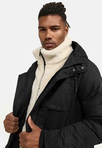 Chaqueta exterior negra con cierre de cremallera, superpuesta sobre un forro polar blanco roto. Presenta un cuello alto y múltiples bolsillos frontales.
