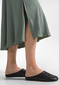 Zwarte instap-muiltjes met een getextureerde afwerking, gecombineerd met een groene, vloeiende jurk met zij-splitten en een afgeronde zoom.