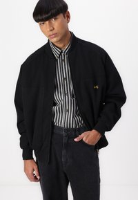 Black bomber jacket with elastic cuffs, front pockets, and a subtle embroidered logo. Paired with a striped button-up shirt and dark jeans.