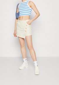 Jupe wrap beige clair avec des boutons en bois, associée à un crop top ajusté rayé bleu et blanc. Le mannequin porte des baskets blanches et des chaussettes.