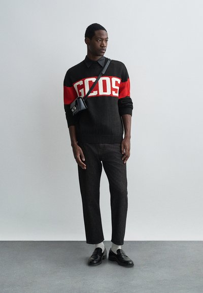 Sweater preto e vermelho com o logótipo "GCDS", jeans escuros, sapatos pretos e uma pequena mala tiracolo preta. Tecido de malha texturizado, ajuste casual.