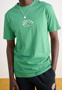 Camiseta verde de manga corta hecha de algodón, con un logo bordado en blanco de "ellesse" y un diseño curvado sutil.