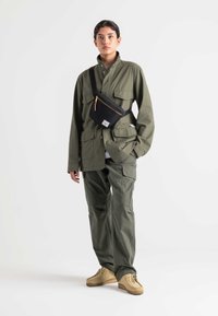 Olive green utility jacket and cargo pants, paired with a black crossbody bag featuring a gold zipper. Suede shoes in a light tan color.