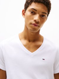 Weißes Baumwoll-V-Ausschnitt-T-Shirt mit einem kleinen gestickten Logo in Rot, Weiß und Blau auf der linken Brust. Glatte Textur, kurze Ärmel.