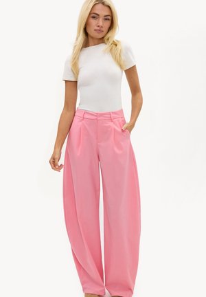 Femme blonde portant un top blanc ajusté à manches courtes et un pantalon rose taille haute à jambes larges, les mains dans les poches.