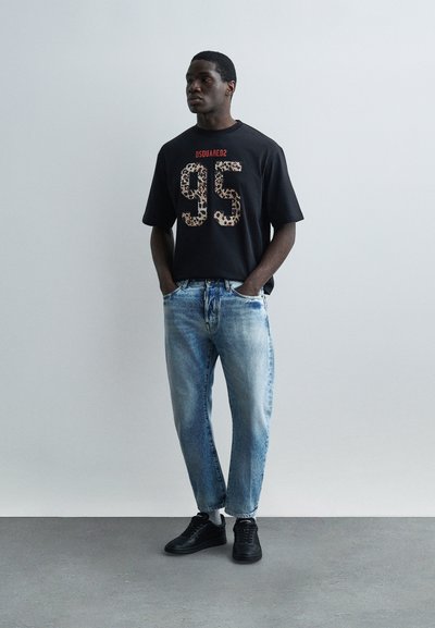 Άνδρας που φοράει μαύρο T-shirt με το κείμενο "95" και λεοπάρ μοτίβο, ανοιχτόχρωμο μπλε τζιν και μαύρα αθλητικά παπούτσια, στέκεται με τα χέρια στις τσέπες.