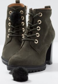 Bottines en daim vert olive à lacets avec talons épais et pompons en fausse fourrure noire sur les lacets, un pompon détaché sur une surface blanche.