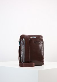 Borsa a tracolla in pelle marrone con una consistenza liscia, dotata di una tasca frontale con cerniera e accenti blu e di una tracolla regolabile.