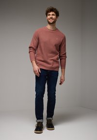Lächelnder Mann steht, trägt einen staubigen rosa Pullover, dunkelblaue Jeans mit Umschlägen und schwarze Schnürstiefel vor einem schlichten grauen Hintergrund.