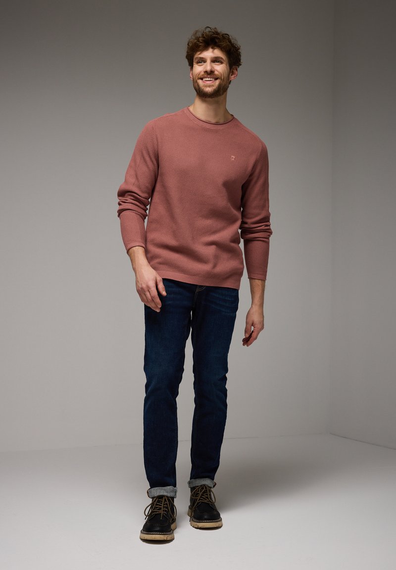 Lächelnder Mann steht, trägt einen staubigen rosa Pullover, dunkelblaue Jeans mit Umschlägen und schwarze Schnürstiefel vor einem schlichten grauen Hintergrund.