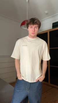 T-shirt beige a maniche corte con la parola 'EGITTO' stampata in nero, abbinato a jeans di denim scuro. Ambientazione interna con un mobile.