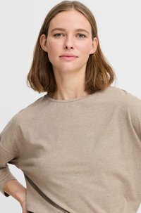 Šviesiai smėlinio atspalvio ilgomis rankovėmis marškiniai su apvalia iškirpte ir švelniu, glotniu audiniu. Modelis pozuoja su viena ranka ant klubų, demonstruodamas laisvą siluetą.