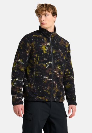 Junger Mann mit lockigem Haar trägt eine schwarze Fleecejacke mit gelben, braunen und weißen Flecken, schaut zur Seite und steht vor weißem Hintergrund.