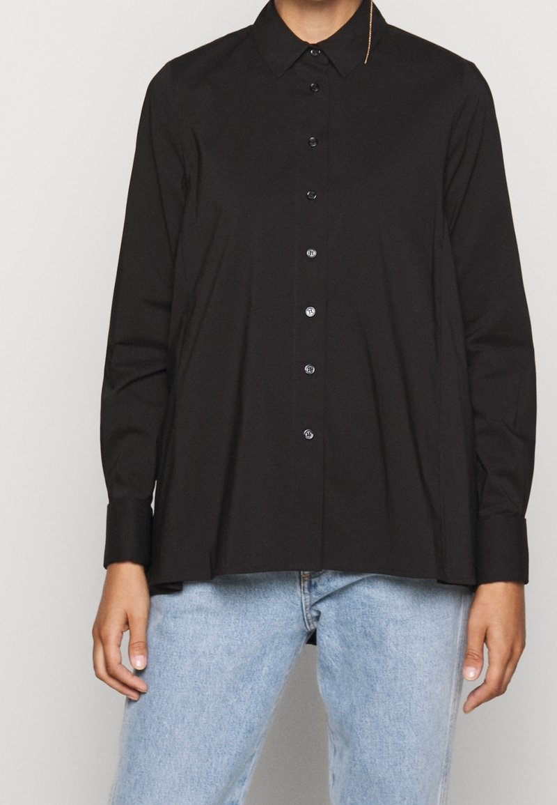 Personne portant une chemise noire à manches longues avec col et boutons, associée à un jean bleu clair, debout devant un fond uni.