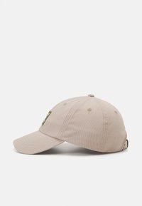 Beige bomull baseball cap med buet skjerm, justerbar stropp og en grønn logo-patch på fronten. Har sydd detaljer på skjermen.