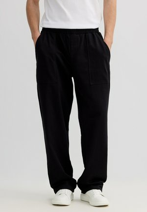 Persona con pantalones negros holgados con bolsillos delanteros de parche, zapatillas blancas y camiseta blanca, con las manos en los bolsillos, de pie frente a un fondo liso.