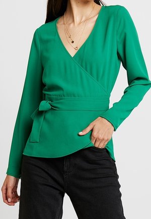 Top verde avvolgente con maniche lunghe, scollatura a V, decorative con un nodo in vita e una texture liscia. Abbinato a pantaloni neri.