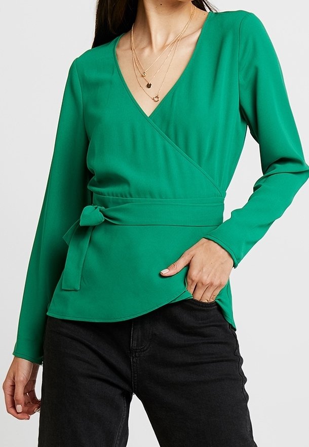 Grünes Wickeloberteil mit langen Ärmeln, V-Ausschnitt, dekorativem Bindeband in der Taille und glatter Textur. Kombiniert mit schwarzen Hosen.