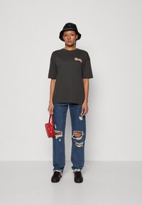 Γκρι oversized T-shirt, μπλε ξεθωριασμένο τζιν, μαύρο καπέλο bucket, κόκκινη τσάντα crossbody, μαύρα αθλητικά παπούτσια με κόκκινες λεπτομέρειες.